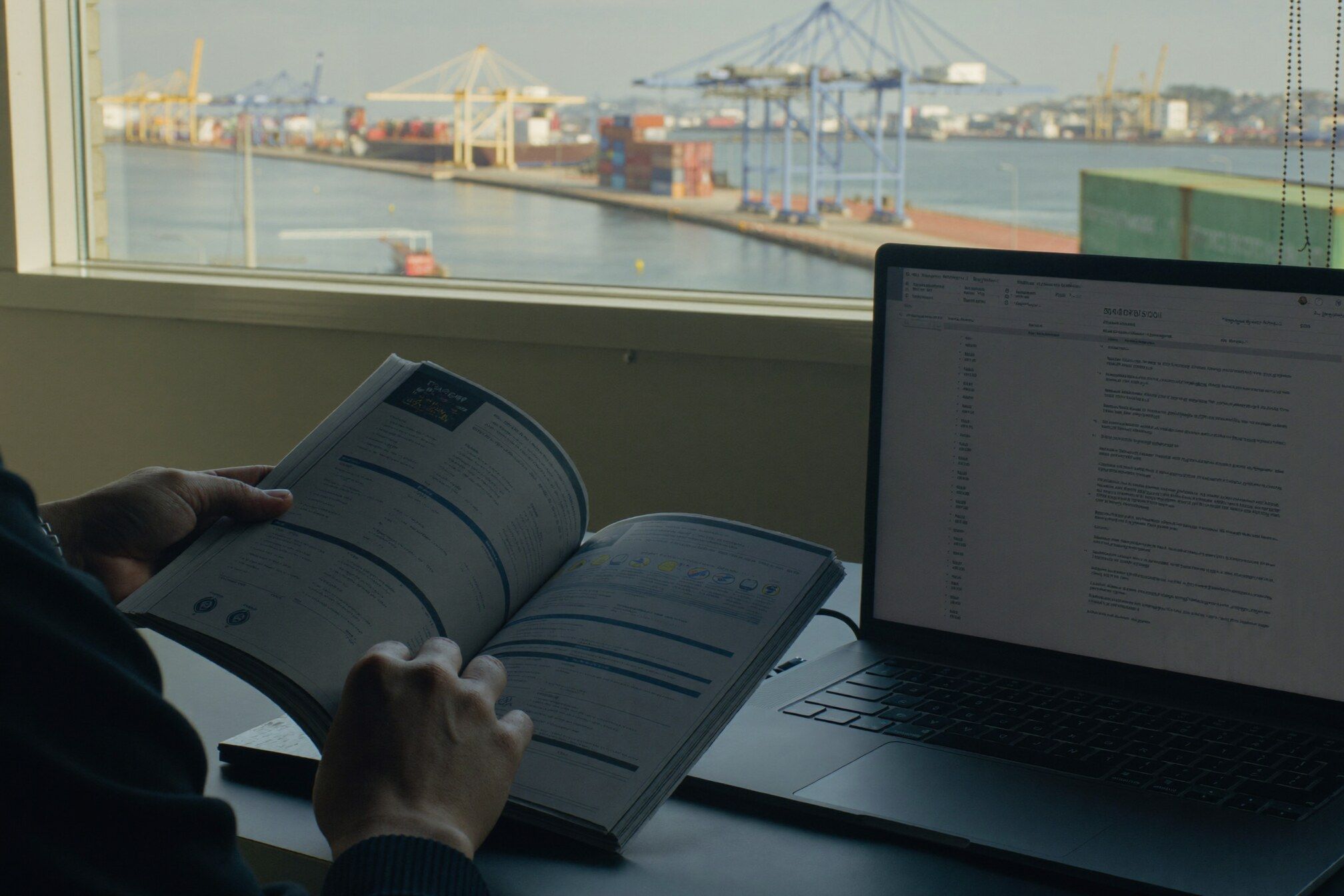 Analista confere a planilha impressa do catálogo de produtos diante do notebook, com o terminal de contêineres na janela